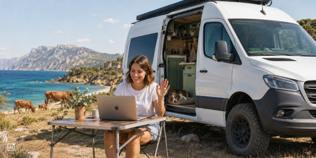 Eine Frau arbeitet vor ihrem Camper an einem Campingtisch und lächelt während Ihres Meetings. Im Hintergrund Ist Ihr Camper, Ihr Hund und die Landschaft und das Meer Siziliens zu sehen.